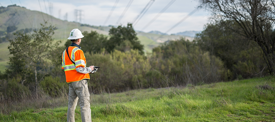 A man in a orange safety vest and white hard hat stands in a right-of-way while practicing environmental stewardship.