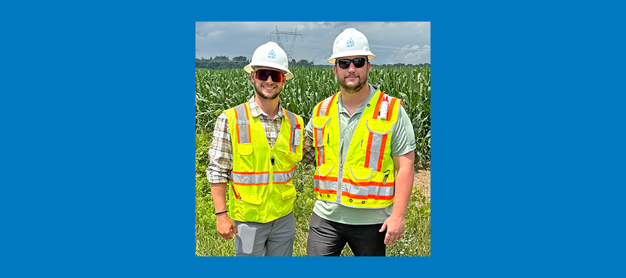 Two brothers wearing white hard hats and high visibility vests in front of a field
