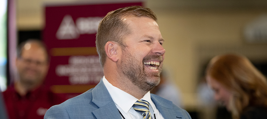 Patrick Geraghty smiling and speaking at a professional event, wearing a blue suit and striped tie, with attendees and an EnviroScience-branded display visible in the background.